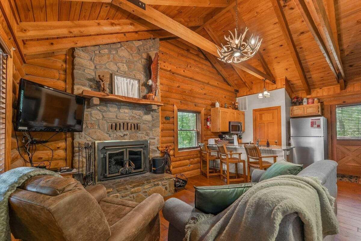Interior de una cabaña de troncos con chimenea de piedra, muebles de madera, televisor y una zona de cocina.