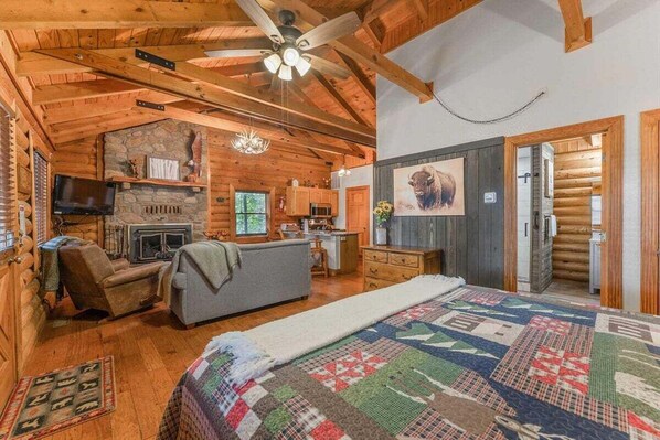 A log cabin interior with a fireplace, a television, a sofa, and a quilted bedspread.