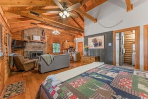 A log cabin interior with a fireplace, a television, a sofa, and a quilted bedspread.