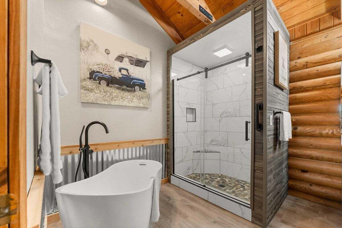 A modern bathroom with a freestanding bathtub, a glass shower enclosure, and a wooden log wall.