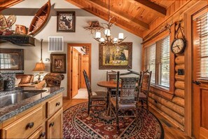 Un interno rustico con soffitto in legno, zona pranzo con sedie a fantasia, cucina con lavello e pensili, e un orologio appeso al muro.