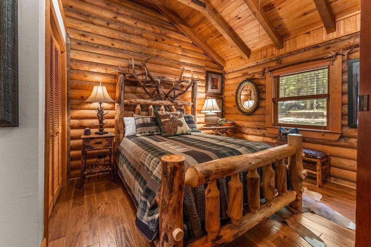 A log cabin bedroom with a four-poster bed, wooden walls, and a window with a view of the outdoors.