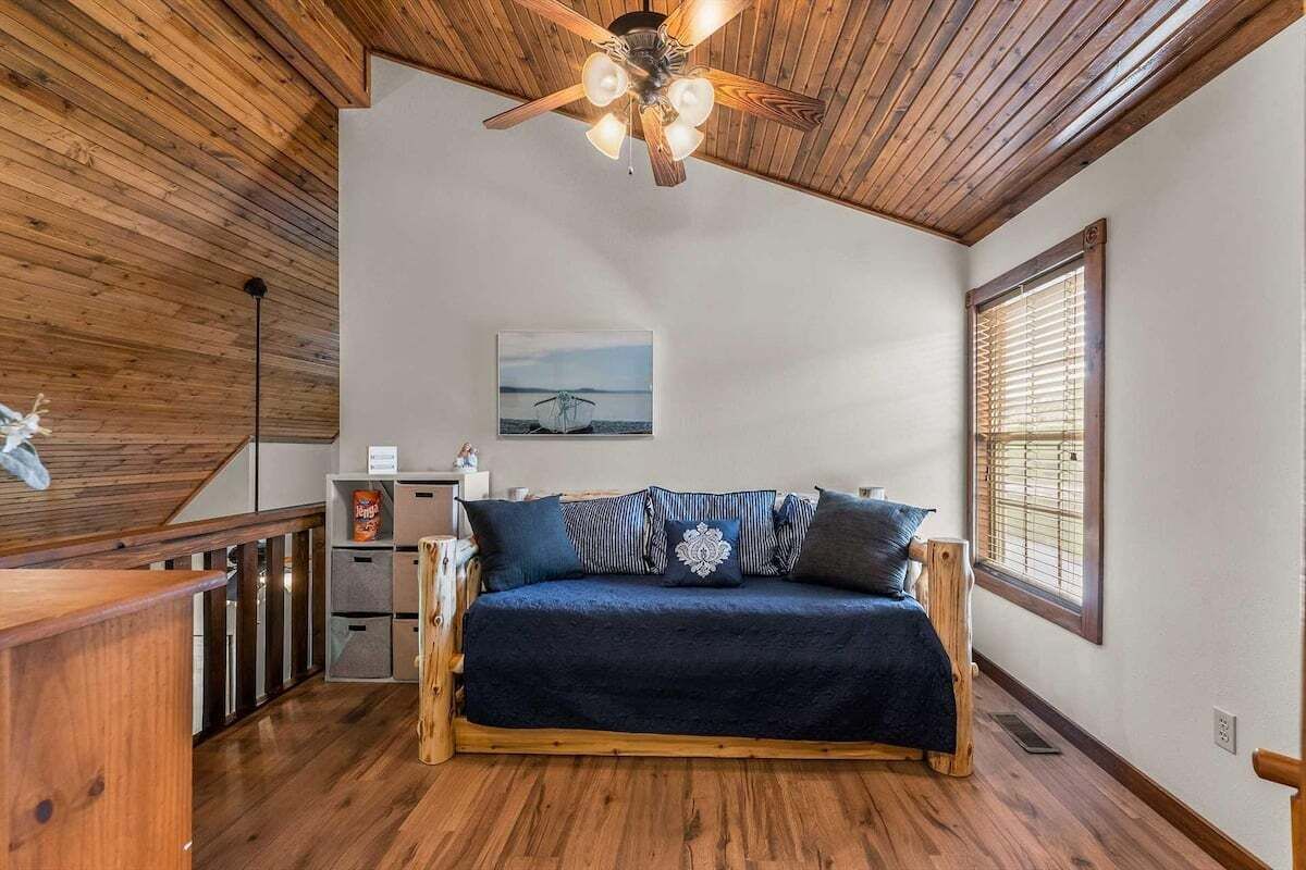 Une pièce avec un plafond en bois, un ventilateur de plafond, un lit recouvert d’une couverture bleu foncé, une fenêtre avec des stores et un tableau accroché au mur.