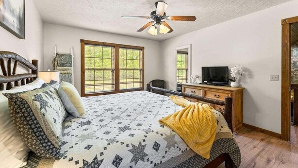 Ein Schlafzimmer mit einem Bett, Deckenventilator, Kommode, Fernseher und einem Fenster mit Blick auf Grünflächen.