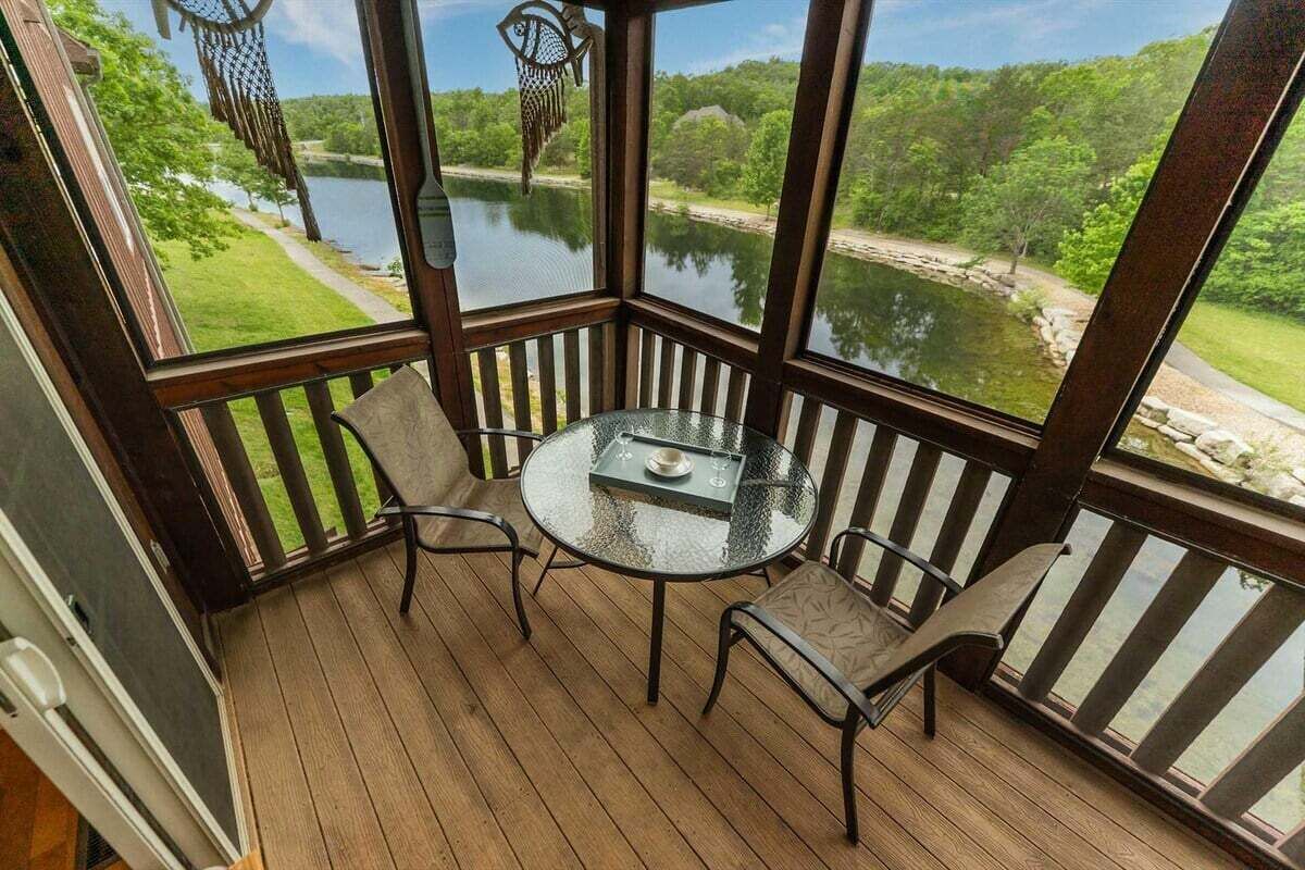 A covered outdoor seating area with a table and chairs, overlooking a body of water and greenery.