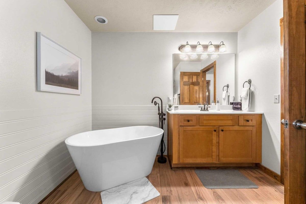 A bathroom with a freestanding bathtub, wooden cabinets, a mirror with lights, and a framed picture on the wall.