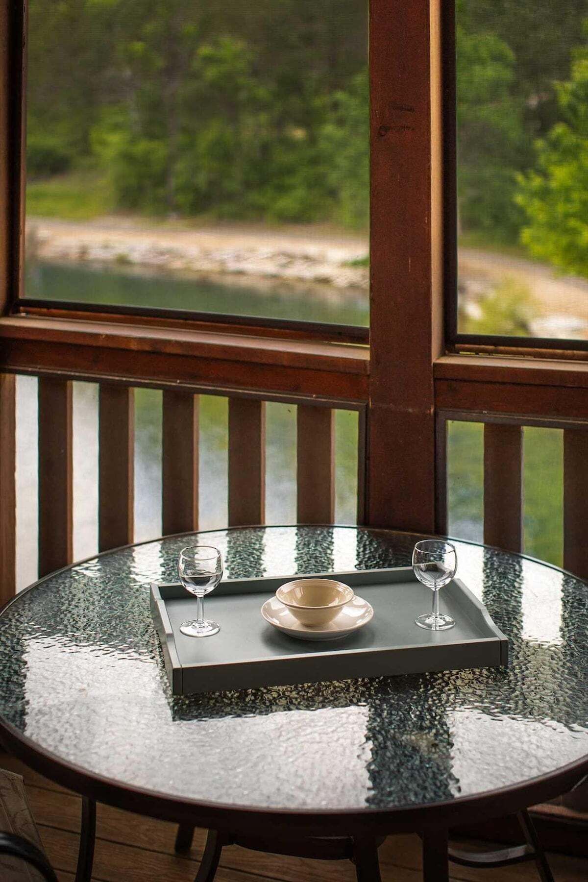 Una mesa redonda de cristal con una bandeja que sostiene un cuenco y dos copas de vino, frente a una ventana con vistas a zonas verdes y un cuerpo de agua.