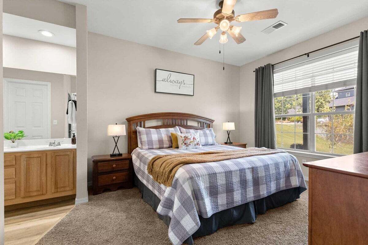Un dormitorio con cama, mesitas de noche, ventilador de techo, ventana con cortinas y baño con lavamanos y armarios.