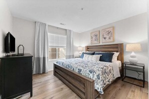 A bedroom with a bed, a bedside table, a lamp, a TV, and a window with curtains. - New Beautifully Renovated Condo w Great Pool View! Free Daily Attraction Tickets (Branson)