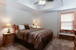 Quarto com cama, duas mesas de cabeceira com luminárias, um banco, ventilador de teto e janela com persianas.