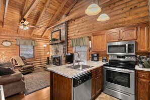 A log cabin interior with a stone fireplace, wooden cabinets, a kitchen with stainless steel appliances, and a cozy seating area.