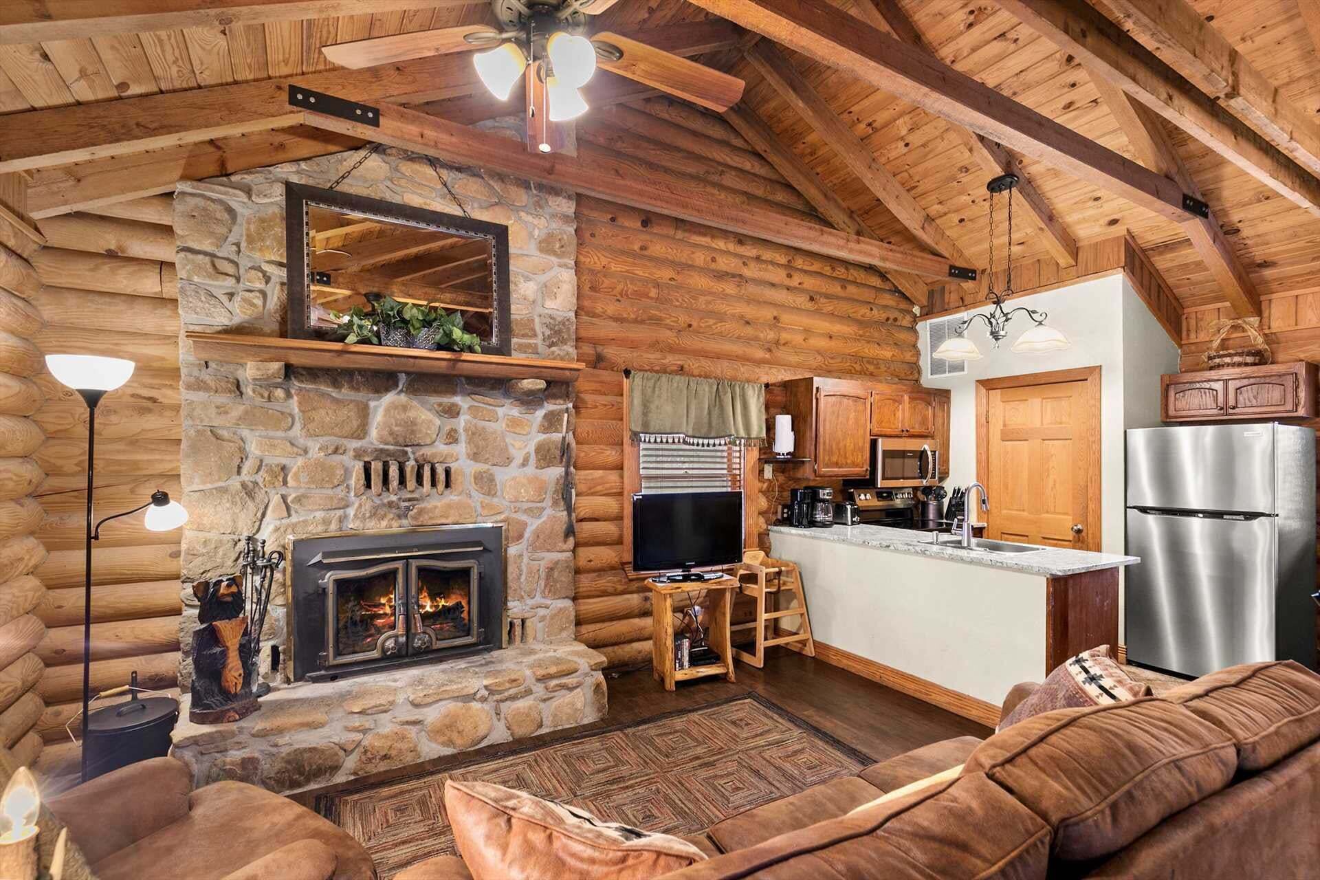 A log cabin interior with a stone fireplace, wooden ceiling, and a kitchenette.