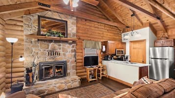 A log cabin interior with a stone fireplace, wooden ceiling, and a kitchenette.