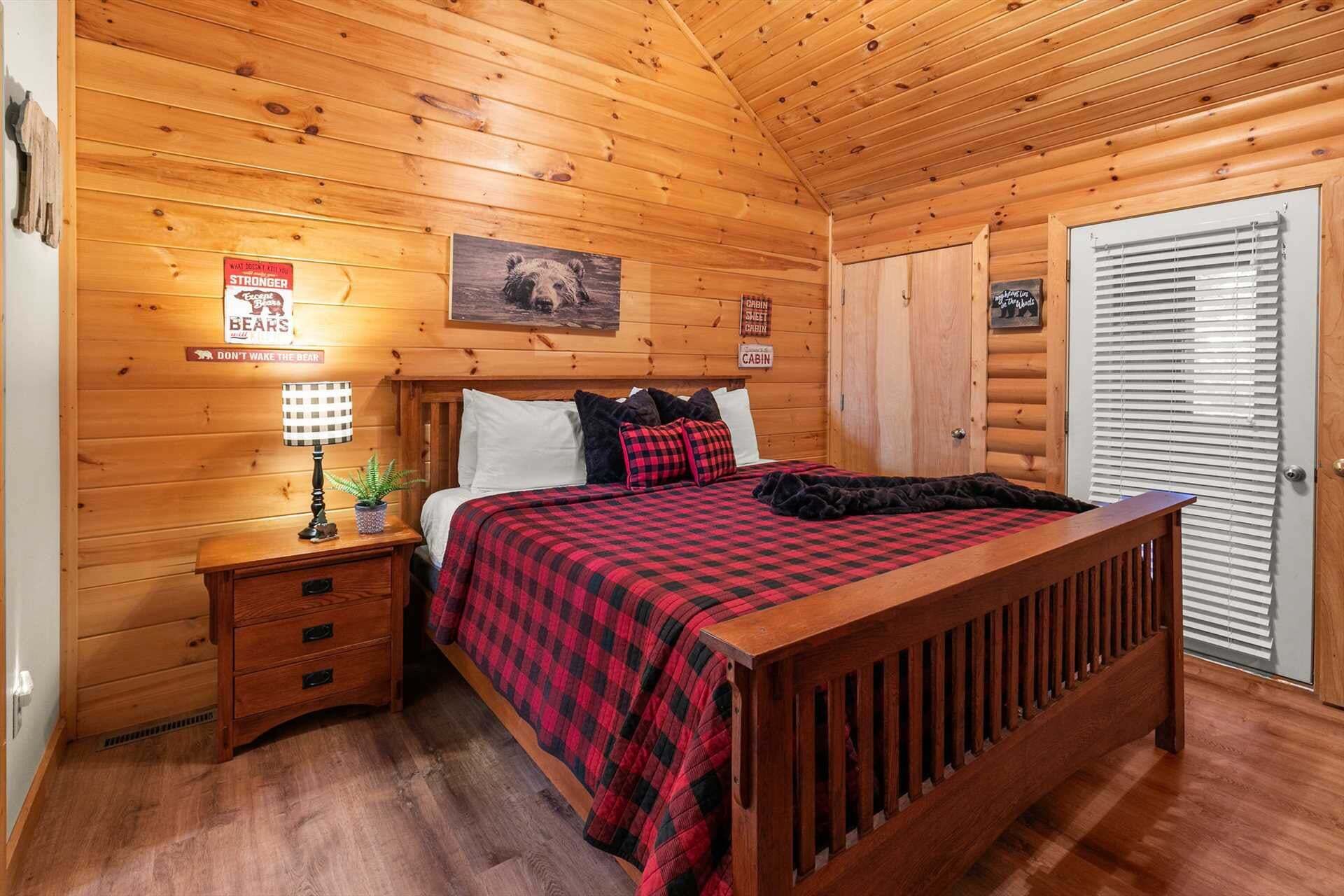 A cabin-style bedroom with a wooden ceiling, a bed with a red and black checkered blanket, a nightstand with a lamp, and a door with a window.