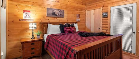 A cabin-style bedroom with a wooden ceiling, a bed with a red and black checkered blanket, a nightstand with a lamp, and a door with a window.