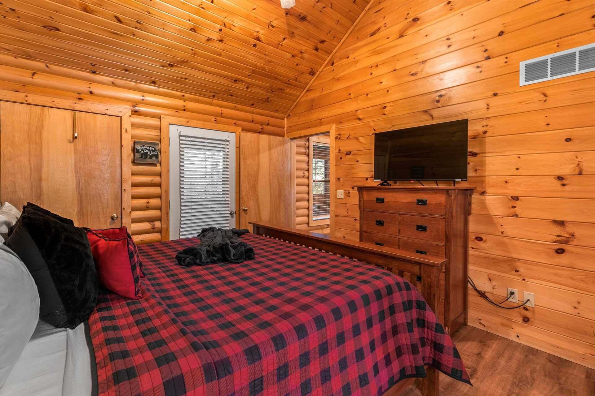 Een slaapkamer in een blokhut met een bed, een dressoir, een televisie en een raam met jaloezieën.