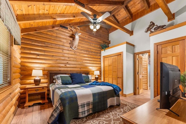 A log cabin bedroom with a bed, nightstands, a ceiling fan, and a television.