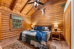 A log cabin bedroom with a bed, two bedside lamps, a ceiling fan, and a window with blinds.
