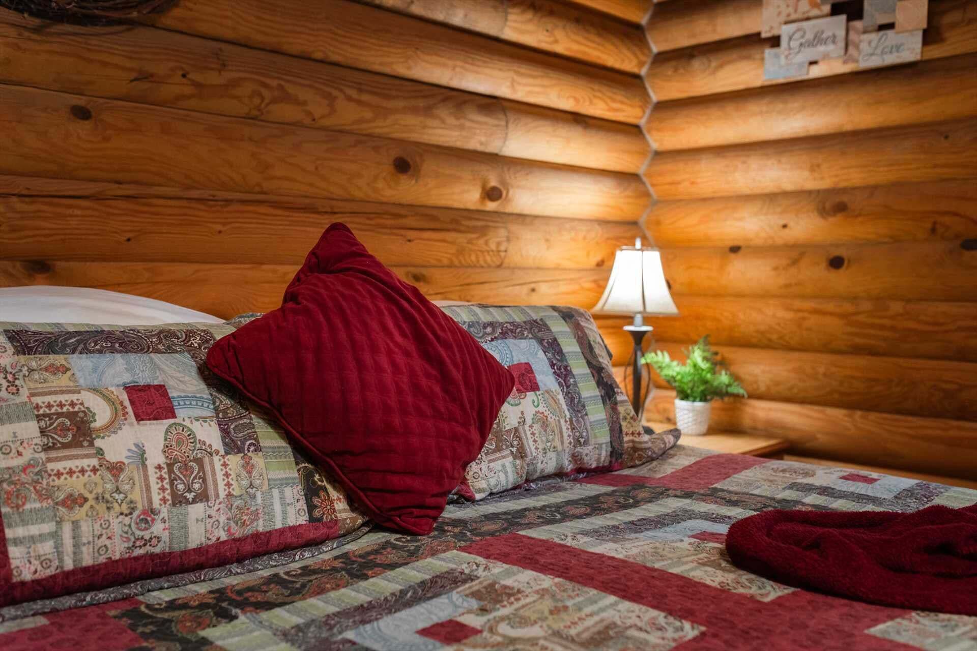 A cozy log cabin-style room with a bed, a red throw blanket, a quilt, a nightstand with a lamp, and a small potted plant.