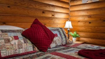 Une pièce chaleureuse, aménagée dans le style d’un chalet en rondins, avec un lit, un plaid rouge, une courtepointe, une table de chevet avec une lampe et une petite plante en pot.