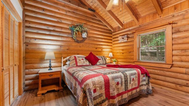 Une chambre à coucher située dans un chalet en rondins, avec un lit, deux tables de chevet, une fenêtre donnant sur un paysage agréable et un ventilateur de plafond.