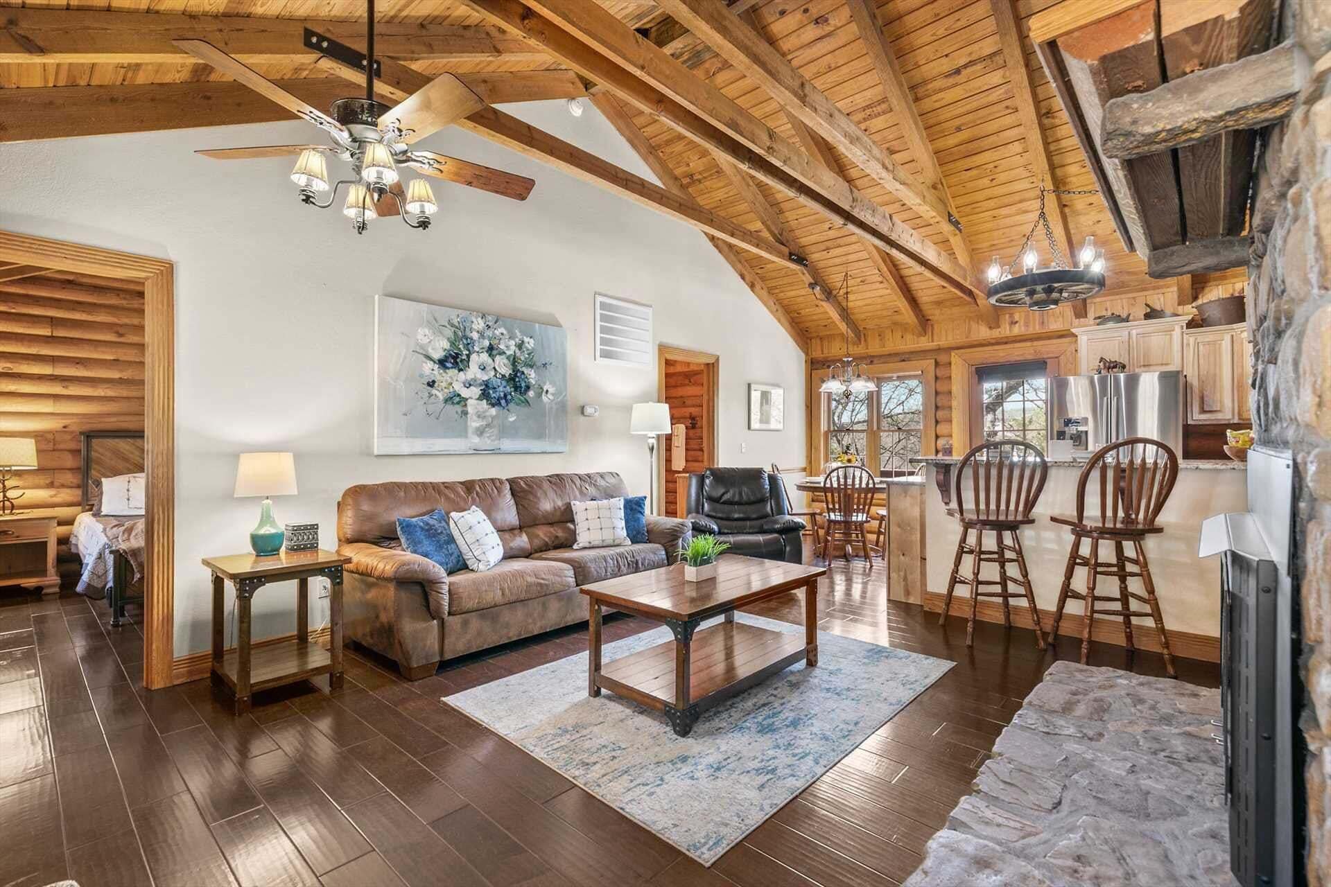 A spacious living room with a high ceiling, wooden beams, a ceiling fan, a fireplace, and a dining area with wooden chairs.