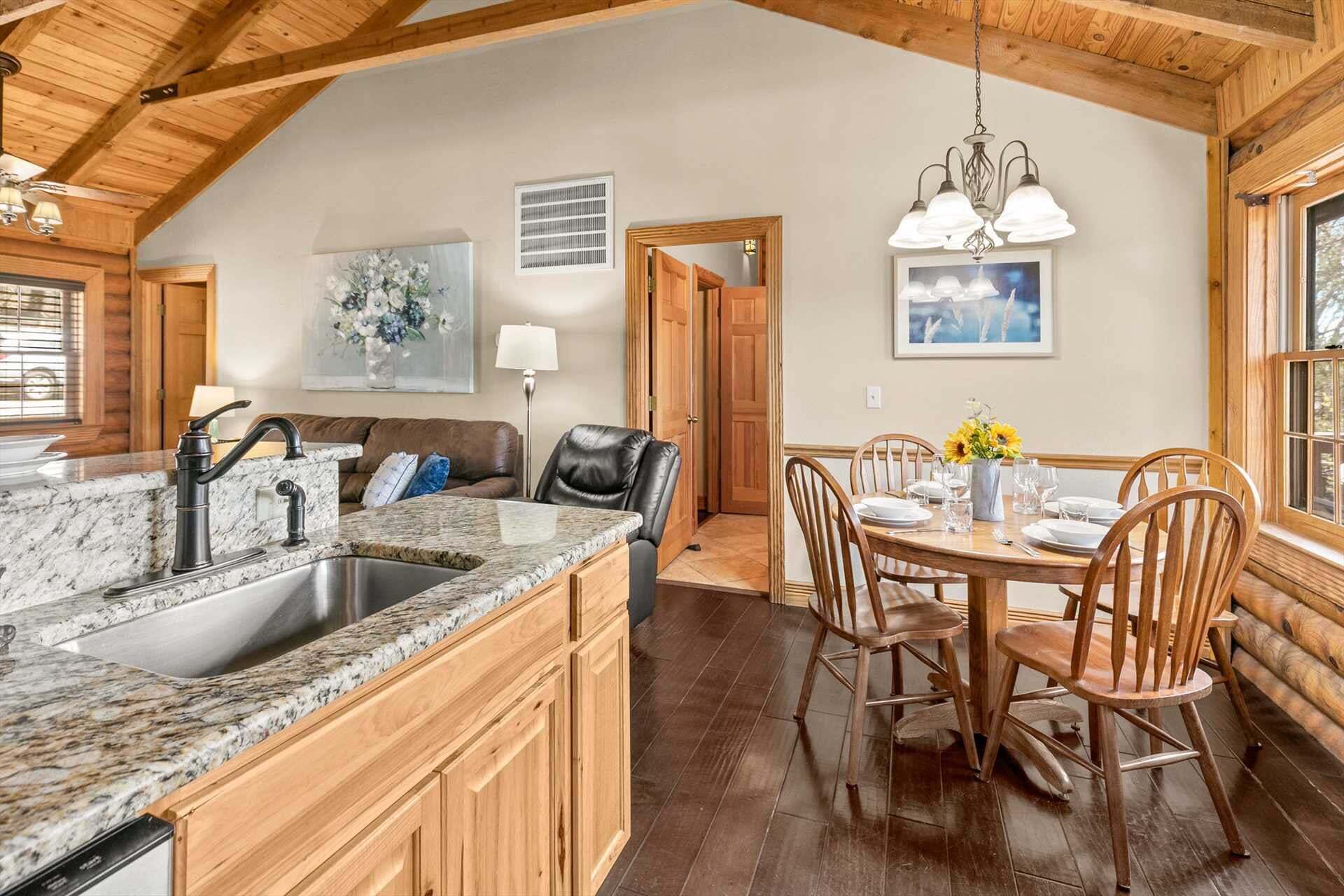 A kitchen with granite countertops, a sink, and wooden cabinets. There is a dining area with a table set for four, surrounded by wooden chairs.