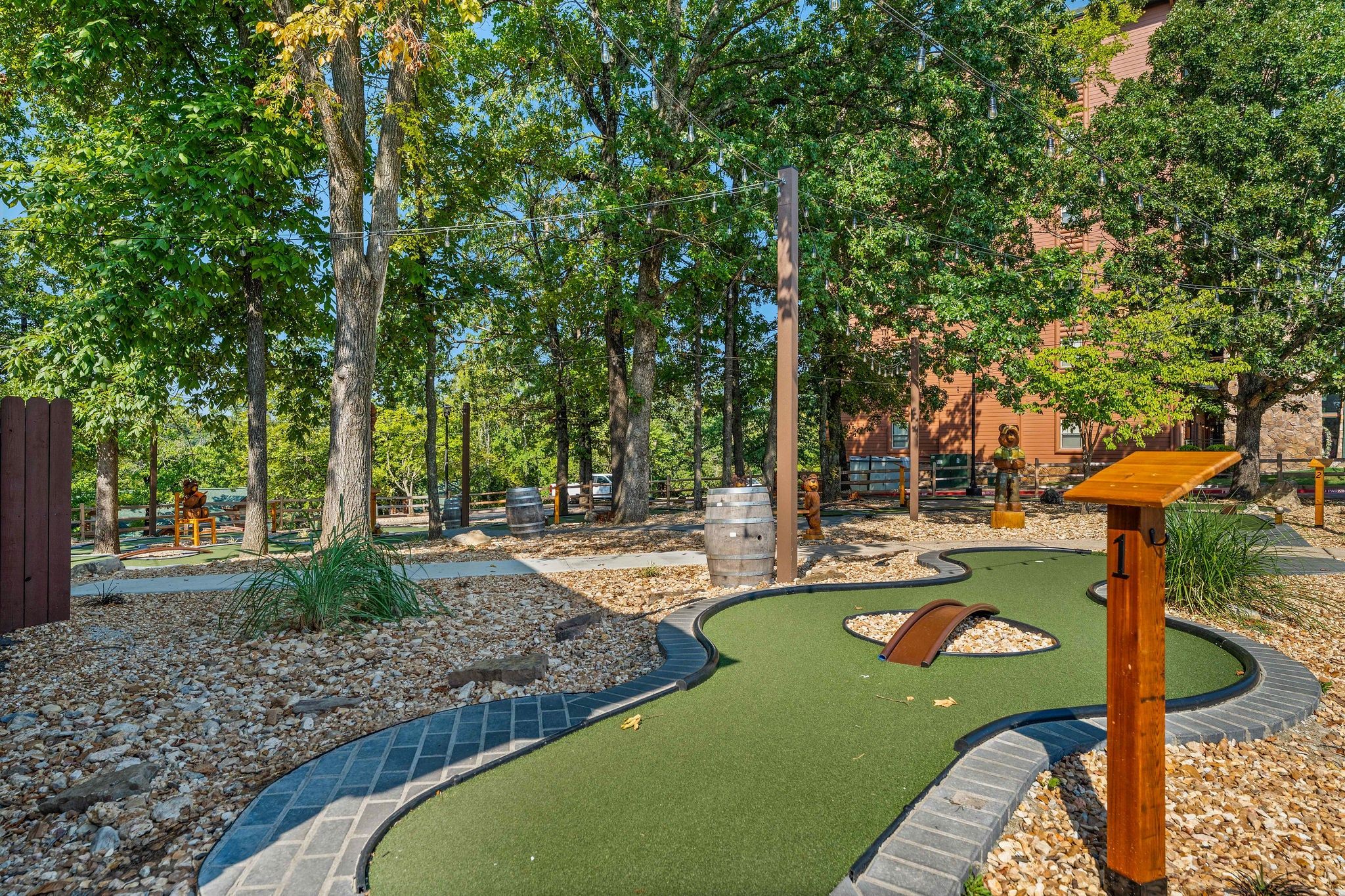 A miniature golf course with a winding path, wooden structures, and a backdrop of trees and buildings.
