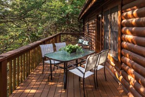 Una terraza de madera con una mesa y sillas de vidrio, rodeada de árboles.