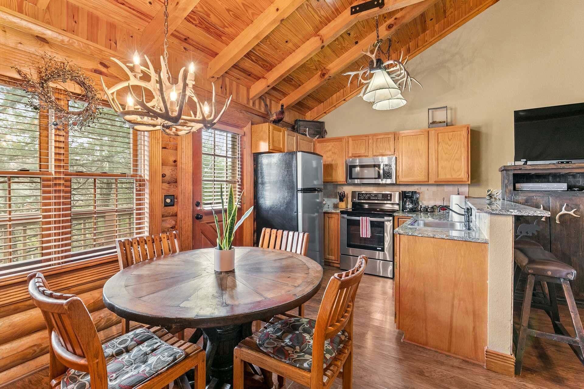 Un intérieur en bois avec une table de salle à manger ronde, des chaises, un lustre et un coin cuisine équipé d’appareils électroménagers.