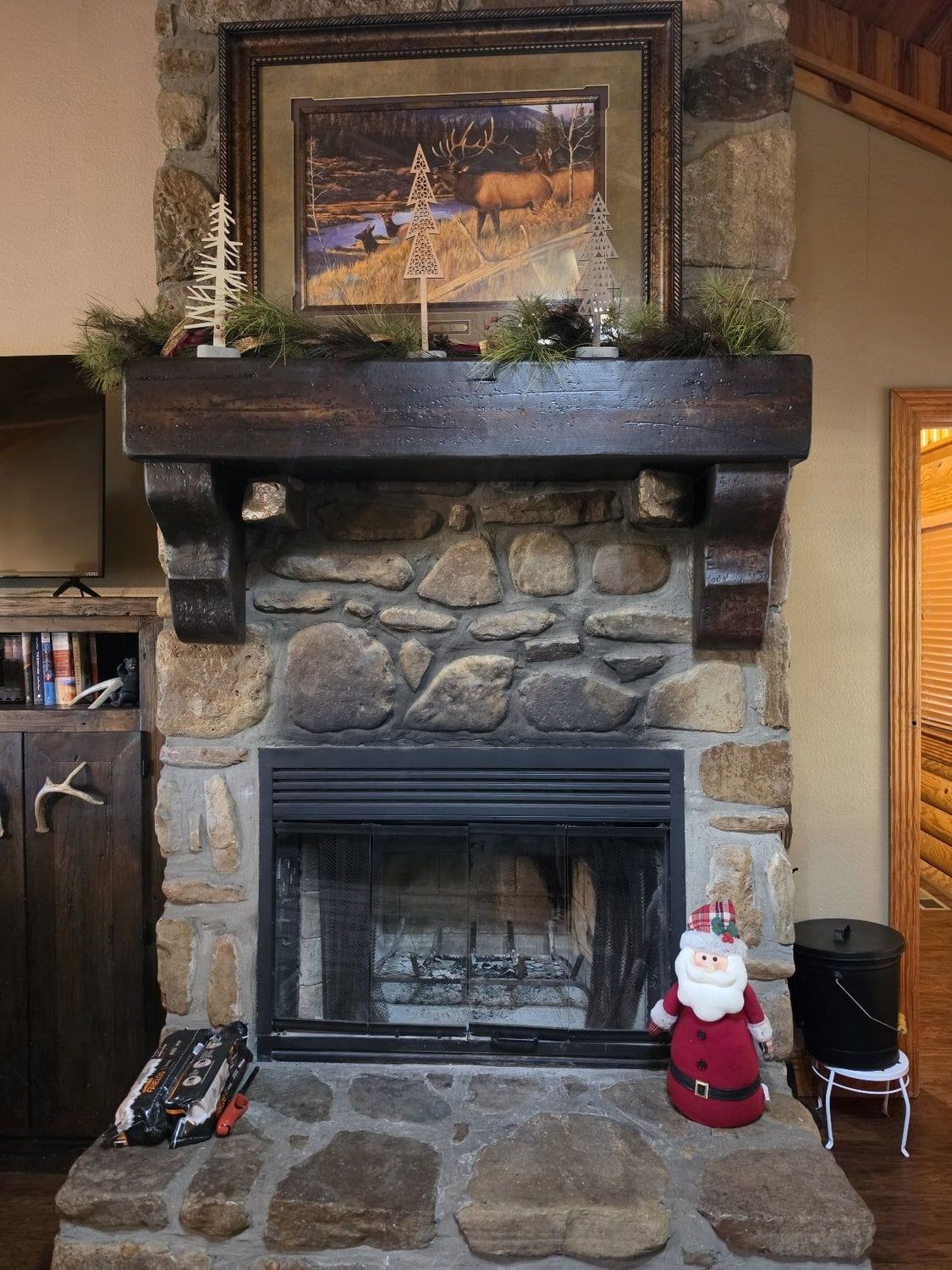 Une cheminée en pierre surmontée d’un tableau encadré, un petit sapin de Noël et une figurine du Père Noël placée à côté.