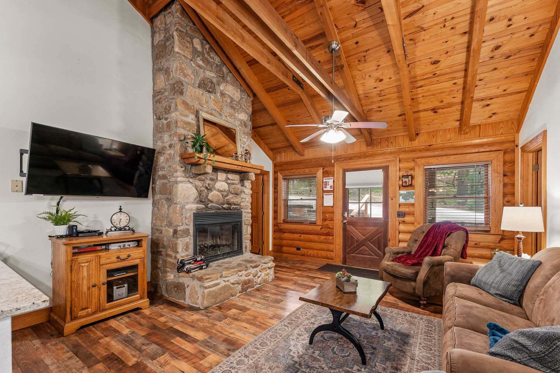 A log cabin interior with a stone fireplace, wooden ceiling, and a television mounted on the wall.