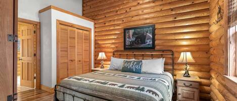 A log cabin bedroom with a bed, bedside lamps, a framed picture on the wall, and a wooden door.