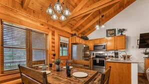 Un intérieur en bois avec une table à manger pour quatre personnes, une cuisine équipée d’appareils électroménagers modernes et une grande fenêtre avec des stores.