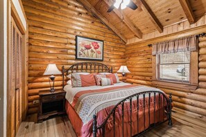 Ein Schlafzimmer in einer Blockhütte mit einem Bett, zwei Nachttischen, einem Fenster mit Jalousien und einem Deckenventilator.