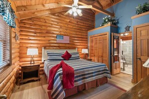 Un dormitorio tipo cabaña de madera con cama, mesita de noche, ventilador de techo y ventana con persianas.