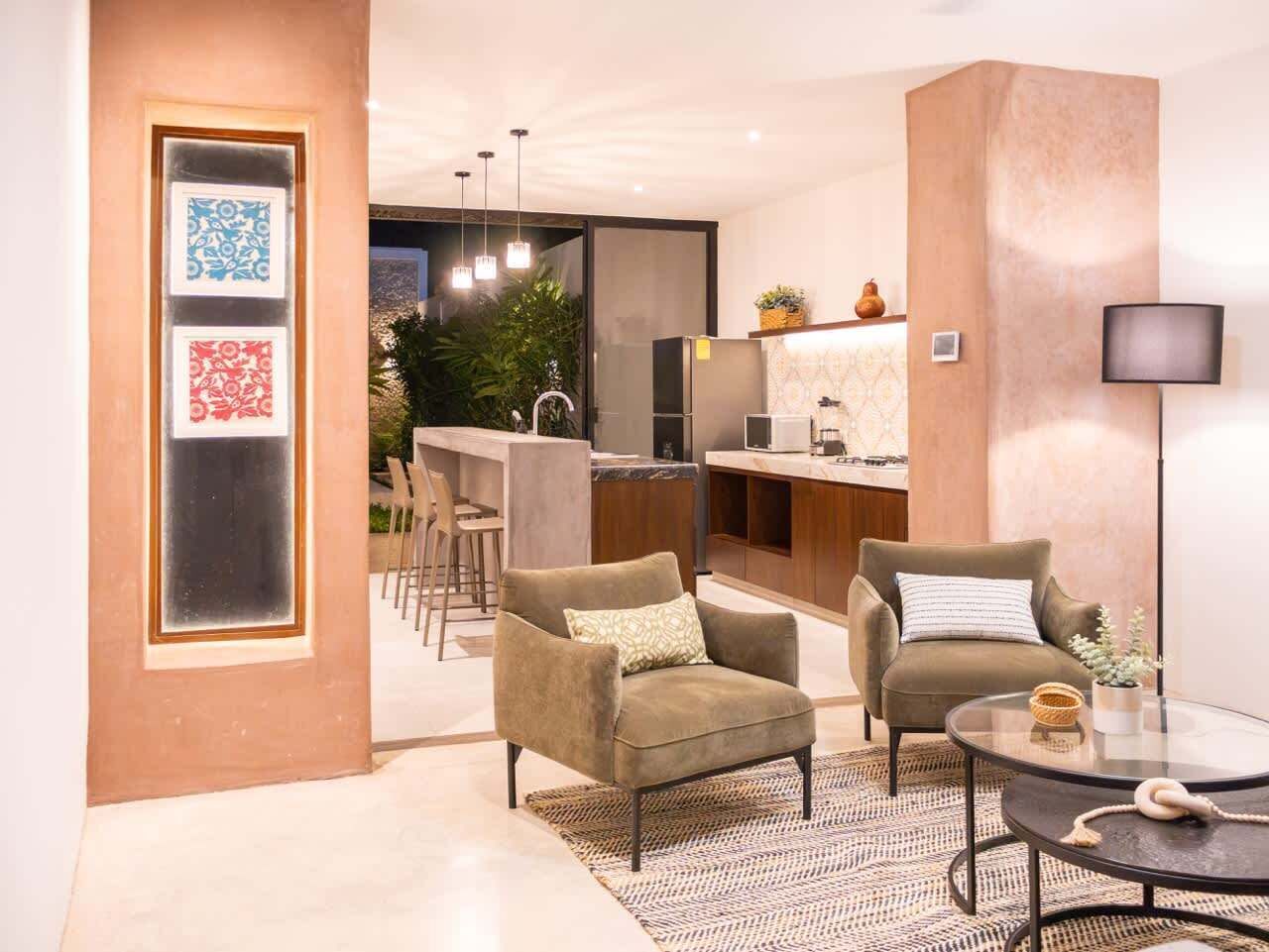 Un salón moderno con dos sillones, una mesa de centro de cristal y una cocina al fondo.