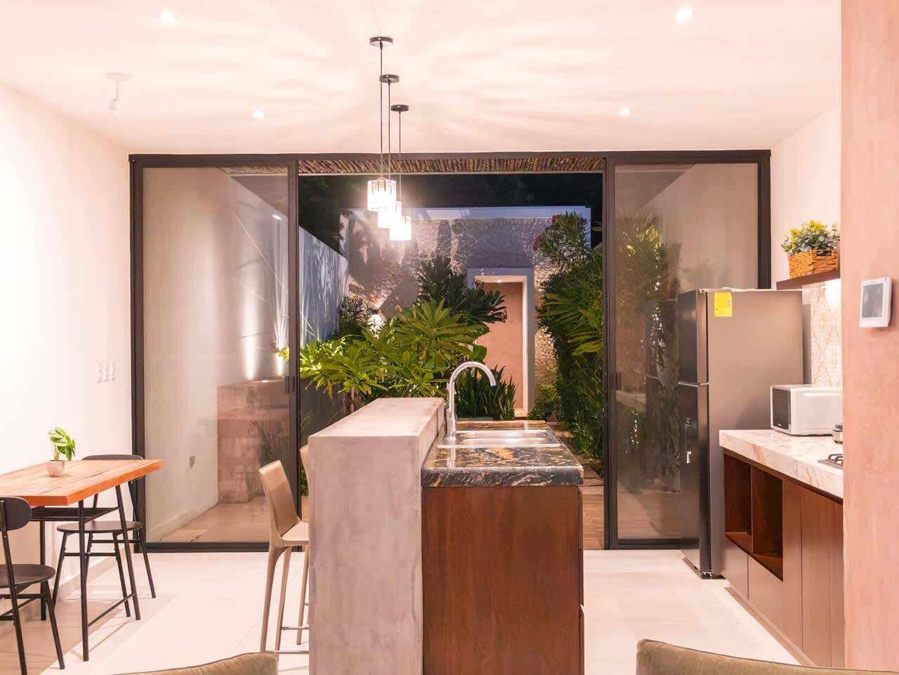 A modern kitchen with a built-in island, a dining area with a table and chairs, and a view of a garden through a glass door.