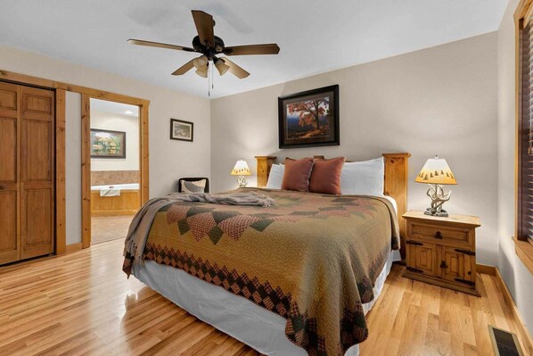 A bedroom with a bed, wooden headboard, two bedside tables with lamps, a ceiling fan, and a door leading to another room.