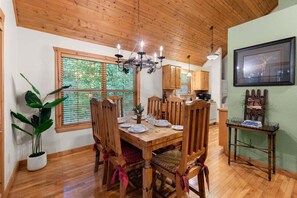 A dining area with a wooden table and chairs, a chandelier, and a view of the outdoors through large windows. - Beautiful Lodge near SDC, Private Fishing Lake, Free Daily Attraction Tickets (Branson West)
