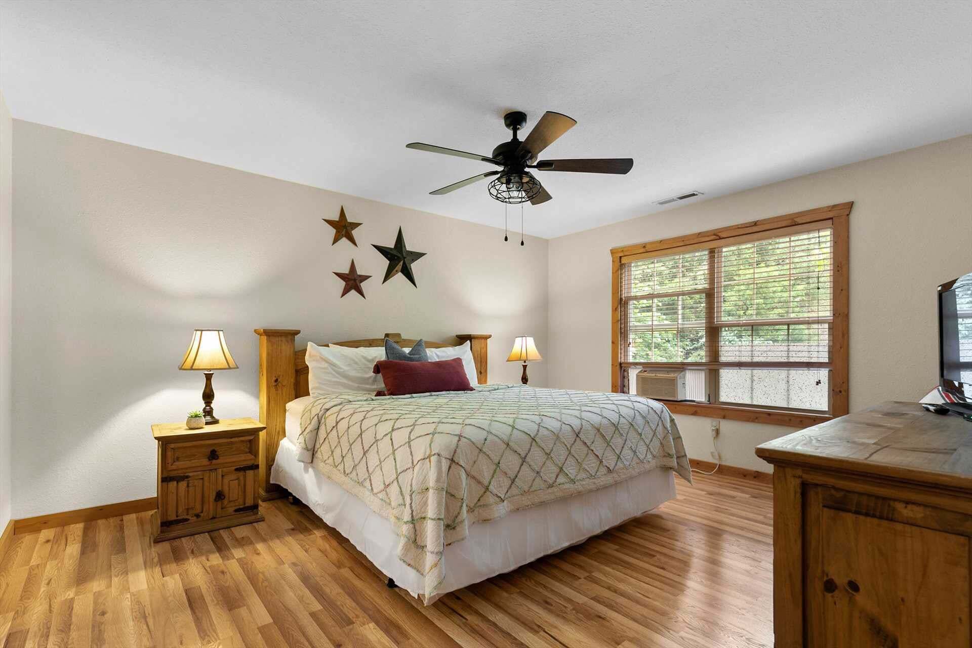 Une chambre à coucher comprenant un lit, deux tables de chevet avec des lampes, un ventilateur de plafond, une fenêtre avec des stores et une commode en bois.