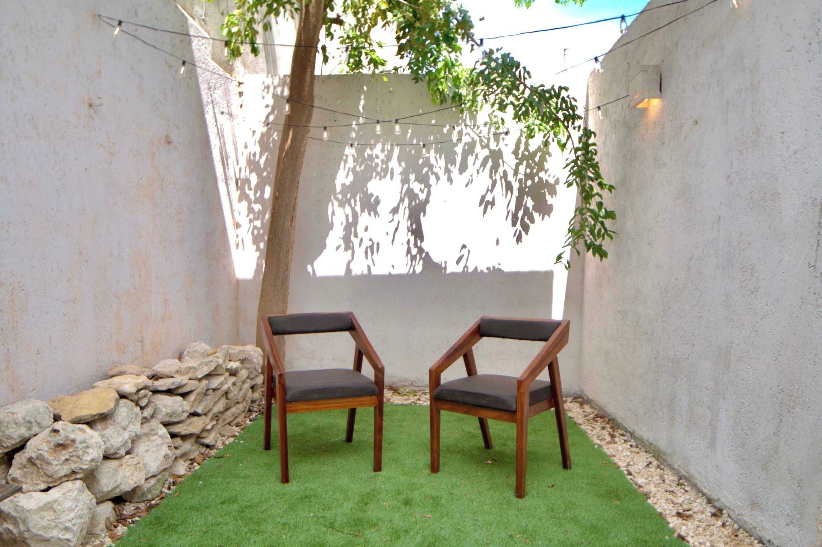 Two wooden chairs with cushions on a green artificial turf, set against a white wall with a stone wall and a tree providing shade.