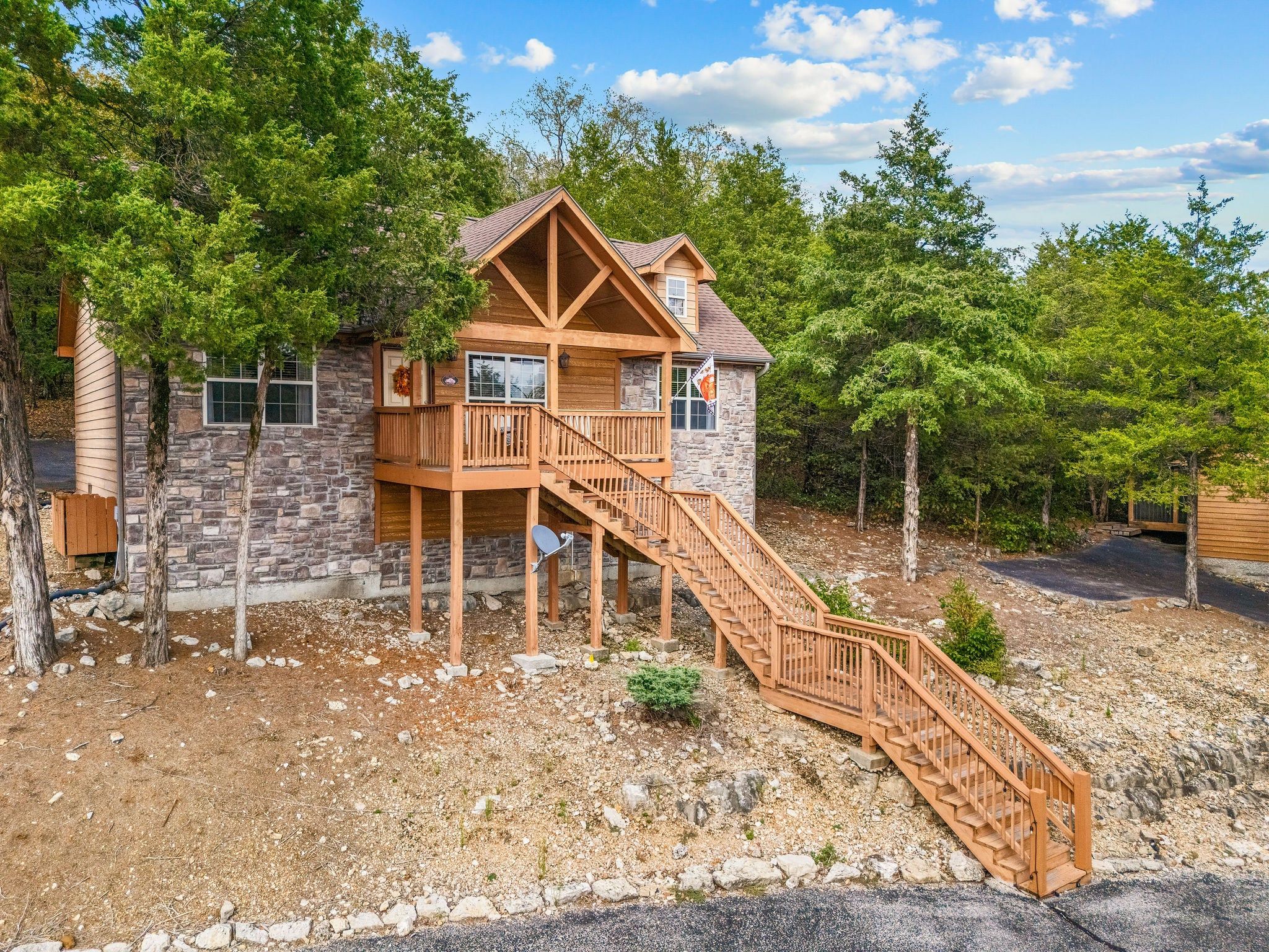 A two-story wooden cabin with a stone foundation, surrounded by trees and a gravel driveway.