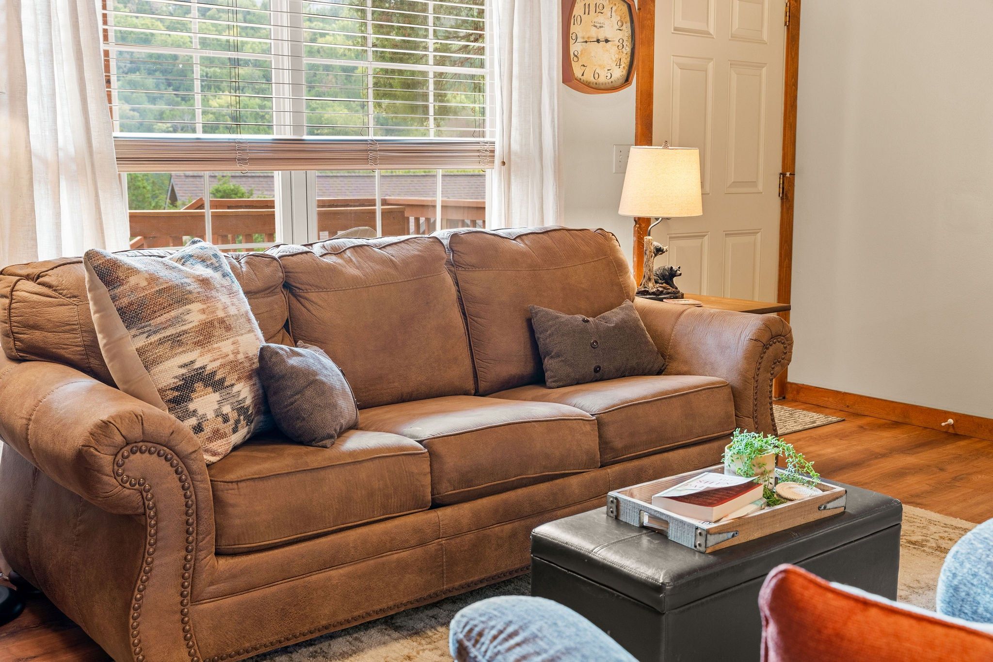 A brown leather sofa with a patterned throw pillow, a small wooden side table with a book and a plant, and a lamp on a wooden nightstand.