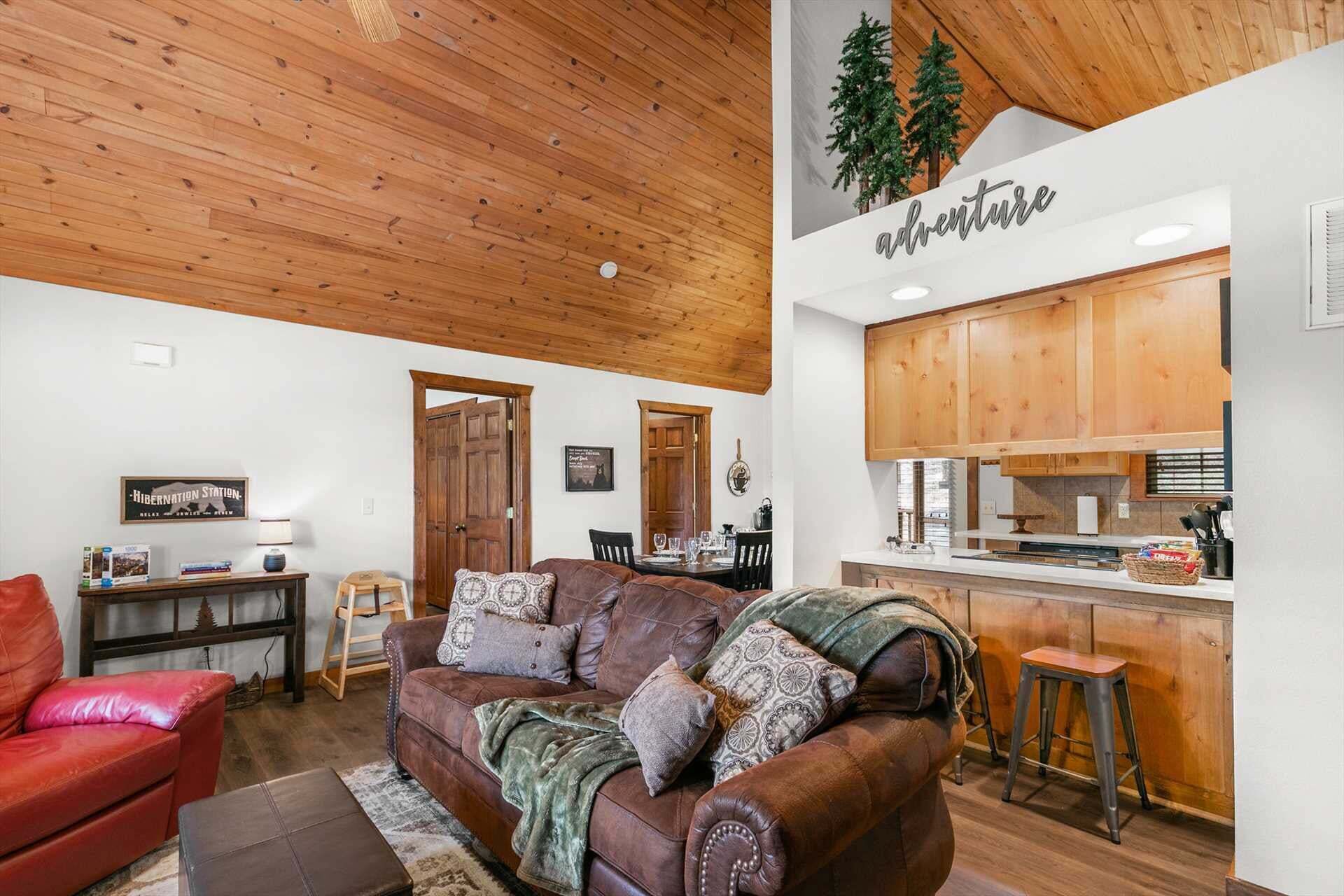 A cozy living room with a brown leather sofa, a red armchair, and a wooden ceiling. A kitchen area is visible with wooden cabinets and a sign that reads 'adventure.'