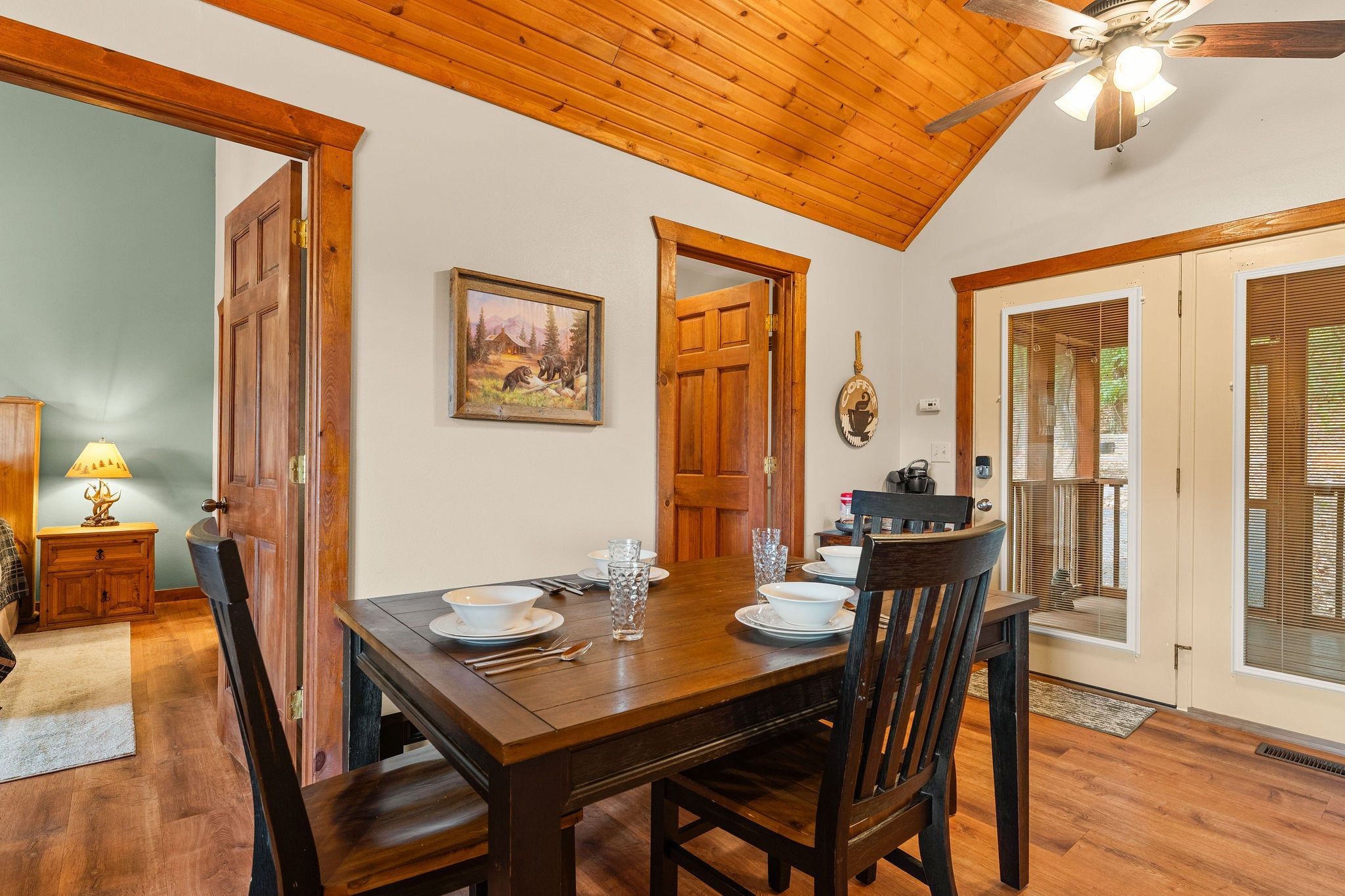 A dining area with a wooden table set for four, a painting on the wall, and a ceiling fan.
