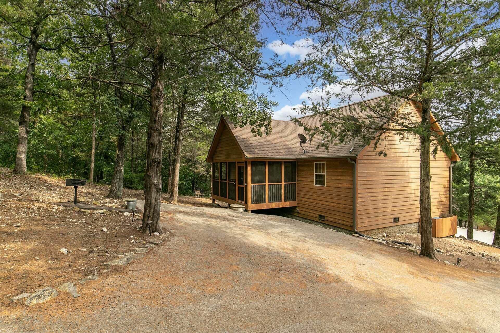 A cabin with a large window, a satellite dish, and a dirt driveway surrounded by trees.
