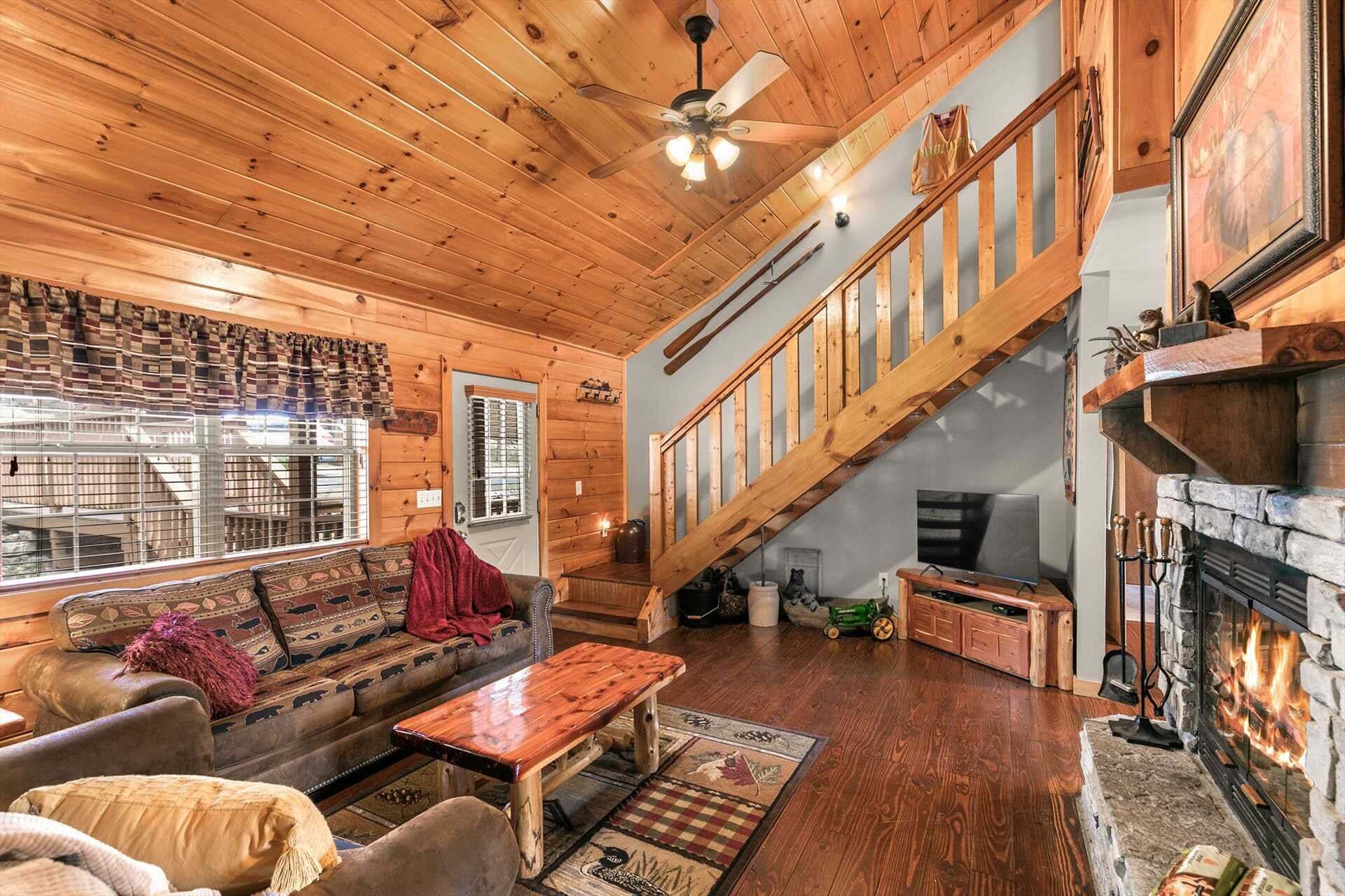 A cozy cabin interior with a wooden ceiling, a fireplace, a staircase, and a living area with a sofa and coffee table.