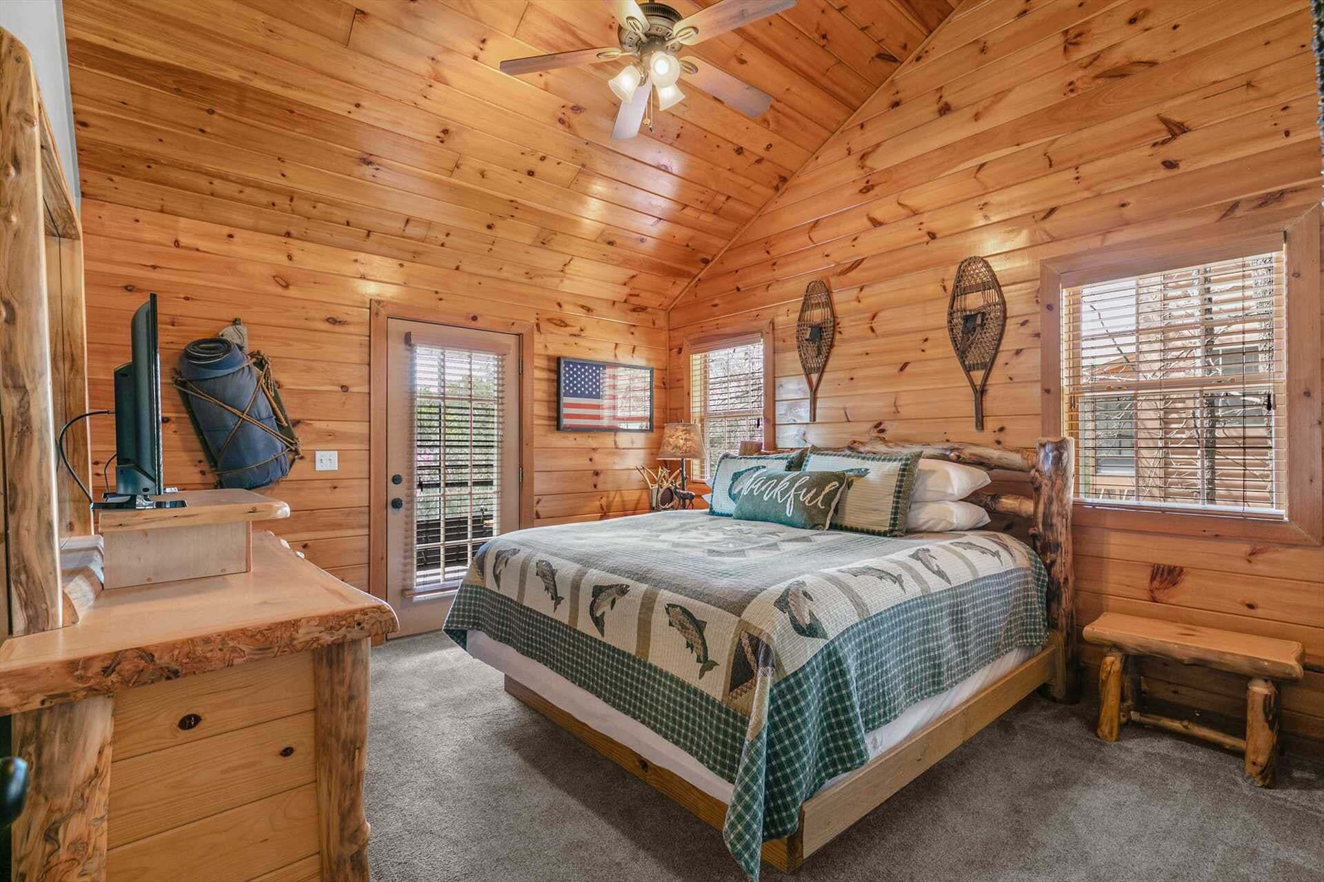 A wooden cabin bedroom with a bed, a desk, a ceiling fan, and a window with blinds.