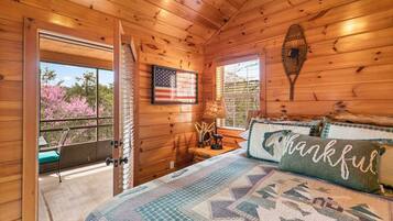 Une chambre dans une cabane en bois, avec un lit, une fenêtre, une porte et une vue sur des arbres aux feuilles roses.